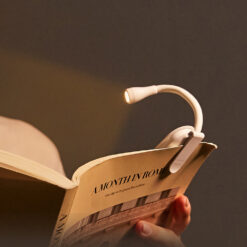 Small black clip-on LED book reading light with a flexible neck attached to a page, illuminating text in a dark room.