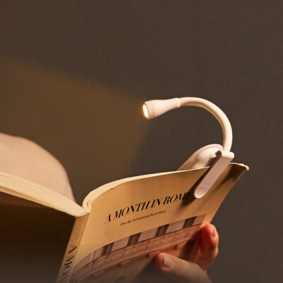 Small black clip-on LED book reading light with a flexible neck attached to a page, illuminating text in a dark room.