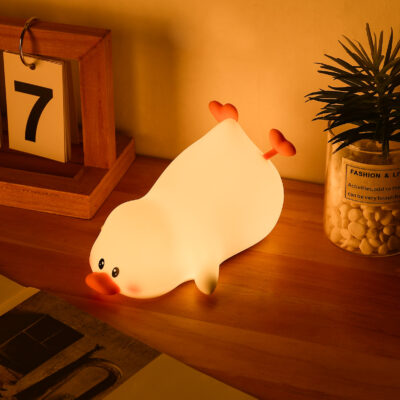 Cute reclining silicone lazy duck night light lamp glowing with a warm, soft light on a bedside table.